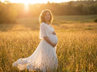 Pregnant woman in a flowing white dress standing in a sunlit grassy field during golden hour.