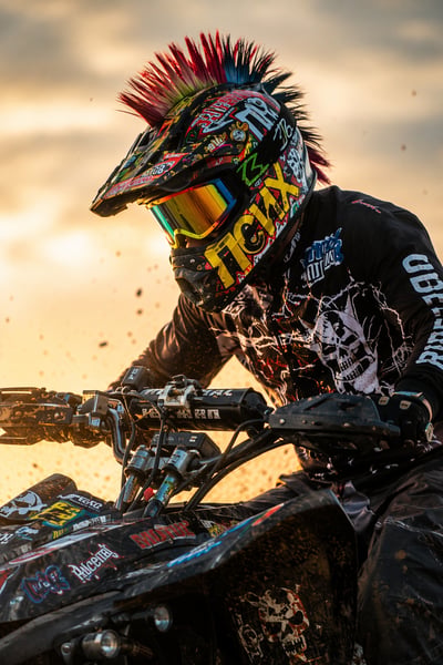 Close-up ATV rider in a graffiti helmet with iridescent visor and mohawk crest, backlit by desert dust at sunset.
