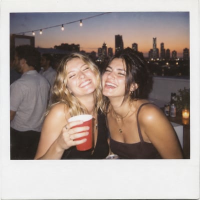 Two friends laughing over a red cup at a rooftop party at dusk, framed by a white instant-film border with string lights, a city skyline, an