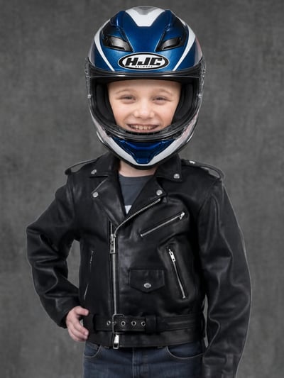 Smiling boy in a blue-and-white HJC helmet, black leather jacket, and dark jeans against a gray studio background
