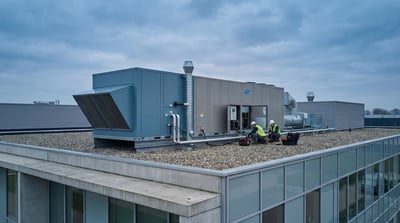 Commercial HVAC rooftop unit with ducting and service workers on a flat building roof under cloudy sky.