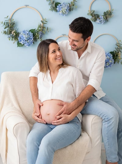 Smiling couple maternity portrait in white outfits with hands on baby bump and soft floral studio decor.