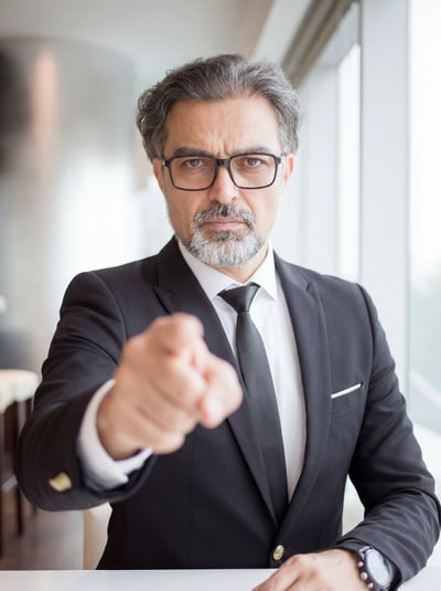 Confident suited business leader pointing toward the camera in a bright office portrait.